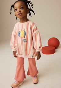 Lyserød stribet sweatshirt med et blåt og gult broderet bille-design, parret med ferskenfarvede flared bukser og lyse sneakers. To runde former i baggrunden.