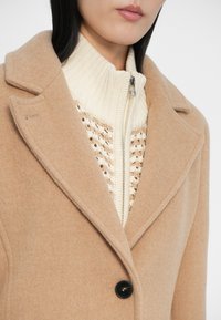 Mujer con un abrigo de lana color camel sobre un suéter de punto crema con cuello alto y cierre, mostrando desde los labios hasta la mitad del torso.
