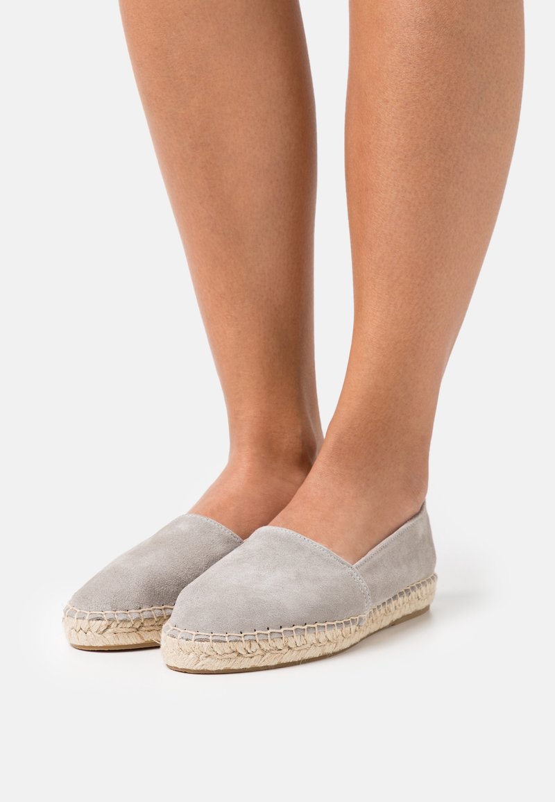 Pieds chaussés d'espadrilles en suède gris clair avec des semelles en jute tressé, posés sur une surface blanche.