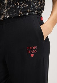 Svarta byxor med resår i midjan och sidoficka, med en röd "JOOP! JEANS"-logotyp och hjärtillustration, tillverkade av ett mjukt, stretchigt material.