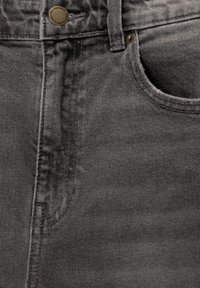 Gros plan sur un jean en denim gris montrant la poche avant, la boucle de ceinture, les coutures et un bouton en métal gravé de la marque.
