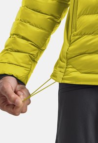 Mano che regola il cordino in vita di una giacca imbottita gialla brillante indossata sopra pantaloni scuri.