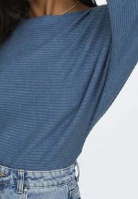 Haut en tissu côtel é bleu avec un col large, des manches drapées et un motif texturé, associé à un jean en denim taille haute.