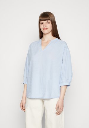 Blusa texturizada azul claro com um decote em V e mangas bufantes de três quartos, feita de um tecido suave. Usada com calças brancas.