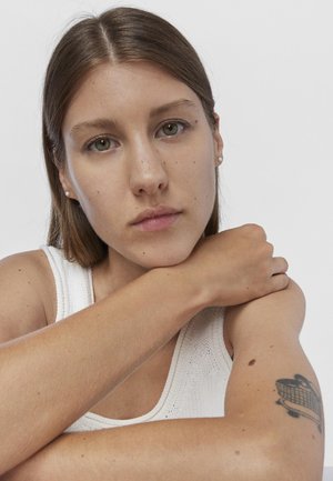 Retrato de una persona con cabello castaño que lleva una camiseta sin mangas blanca y texturizada; sus rasgos incluyen ojos verdes, pecas y un tatuaje visible en el brazo.