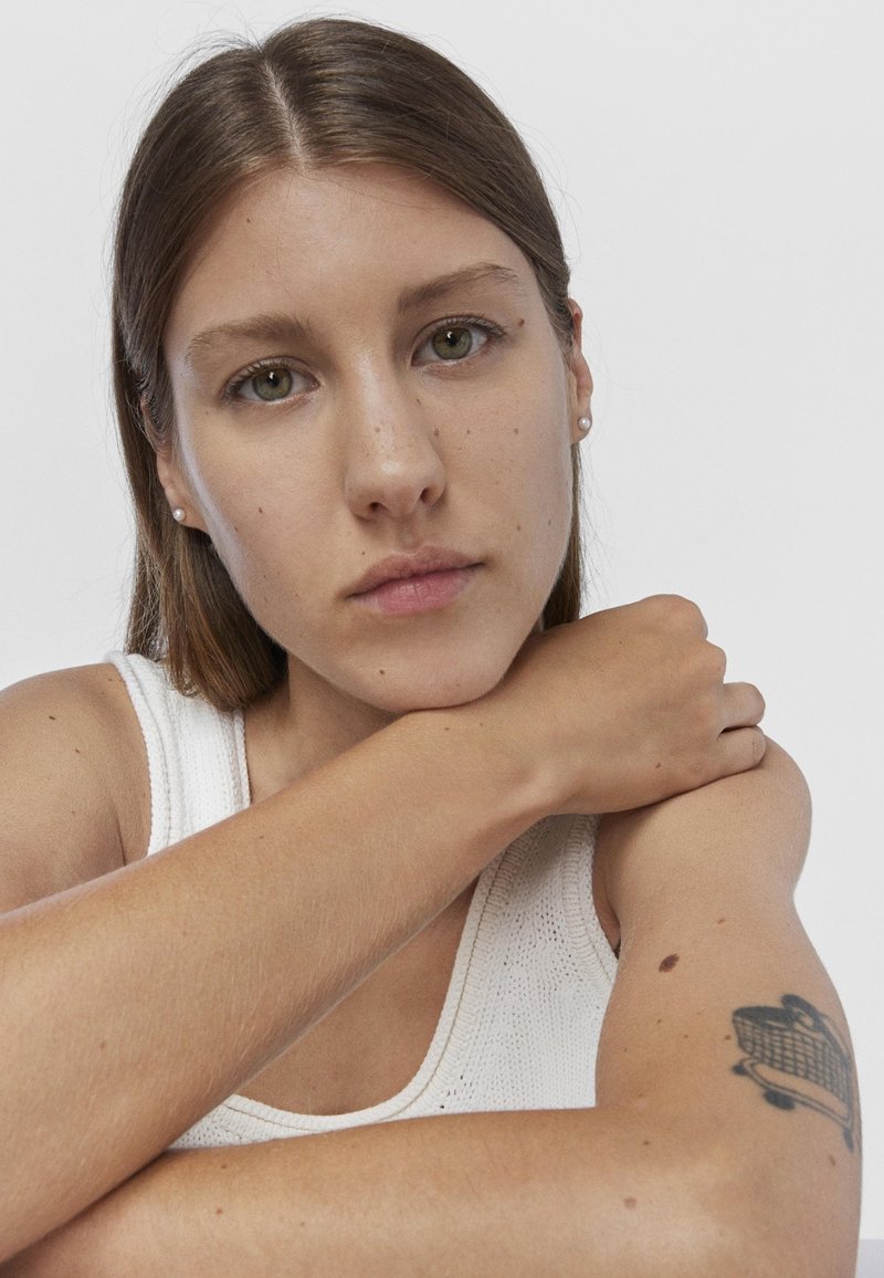 Retrato de una persona con cabello castaño que lleva una camiseta sin mangas blanca y texturizada; sus rasgos incluyen ojos verdes, pecas y un tatuaje visible en el brazo.