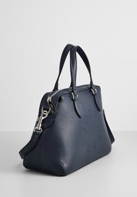 Granatowa skórzana torebka o zaokrąglonym kształcie, z podwójnymi uchwytami i srebrnymi elementami metalowymi. Z boku znajduje się wytłoczona detaliczna logo.