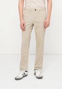 Mann iført beige bukser med rette ben og hvite joggesko med svarte striper, stående mot en enkel hvit bakgrunn.