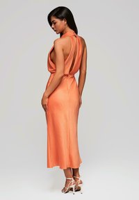Robe en satin orange avec un dos drapé et un design à col halter. Texture lisse, longueur mi-mollet, assortie à des sandales à talons hauts à brides.
