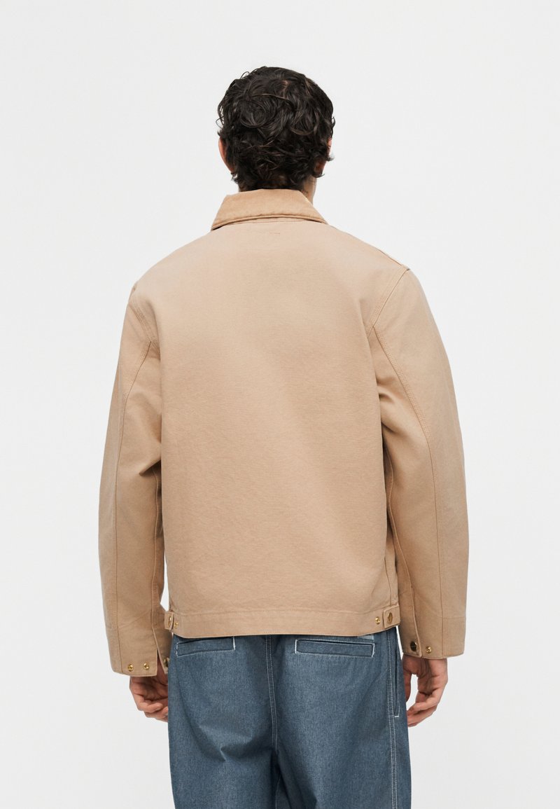 Uomo con capelli ricci che indossa una giacca beige e jeans blu, visto di spalle contro uno sfondo bianco uniforme.