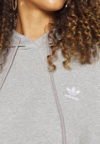 Sweat à capuche gris en matière douce, avec un col en V, des cordons de serrage, et un logo Adidas blanc brodé sur le côté gauche de la poitrine.