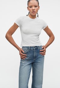 Bela, kratka majica s kratkimi rokavi, ujemajoča se s svetlo modrimi naravnost krojenimi jeans hlačami, ki imajo gumbasto zapiranje in klasičen pet-džepni dizajn.