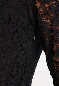 Czarna koronkowa tkanina w kwiatowy wzór z prześwitującymi sekcjami ukazującymi skórę pod spodem, podkreślająca skomplikowane wzory i fakturę w zbliżeniu.