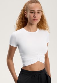 Valkoinen lyhythihainen crop-top, jossa on rypytys yksityiskohtana edessä, valmistettu sileästä, joustavasta kankaasta. Yhdistetty mustiin housuihin.
