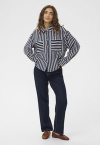 Črtasta majica v mornarsko modri in bež barvi, z gumbi spredaj, dvema žepoma na prsih in dolgimi rokavi. Kombinirana s temnimi denim kavbojkami in rjavimi čevlji.