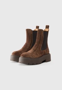 Botas de tobillo de ante marrón con paneles elásticos en los laterales, una suela gruesa y plataforma, y un diseño de tracción robusto. Presentan costuras de contraste y tiradores.
