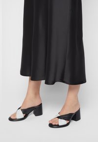 Black and white heeled mules featuring a twisted design, smooth leather texture, and block heel. The mules have an open toe style.
