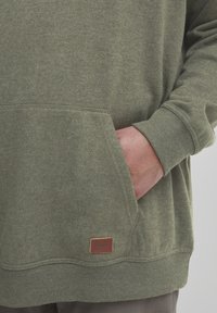 Sudadera verde oliva con un acabado texturizado, presenta un bolsillo tipo canguro y puños acanalados. Una pequeña etiqueta de cuero está unida en el dobladillo.