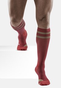 Rote kniebedeckende Socken mit geripptem Design, zwei horizontalen goldenen Streifen oben und verstärkten Zehen- und Fersenbereichen für Haltbarkeit.