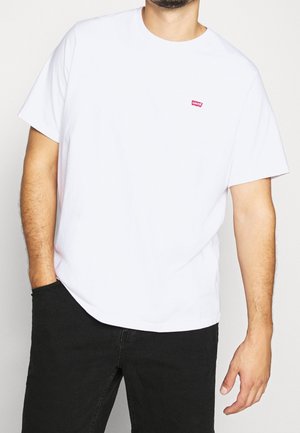 Mann trägt ein schlichtes weißes T-Shirt mit kurzen Ärmeln und kleinem rotem Levi's-Logo auf der Brust sowie schwarze Hose, eine Hand in der Hosentasche.