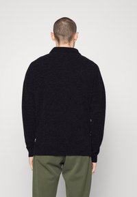 Schwarzer, strukturierten Strickpullover mit Schalkragen und langen Ärmeln, kombiniert mit olivgrünen Hosen. Die Rückansicht hebt Form und Länge hervor.