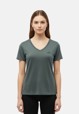 Giovane donna con capelli castani lisci che indossa una maglietta verde opaco a maniche corte con scollo a V, in piedi davanti a uno sfondo chiaro e semplice.