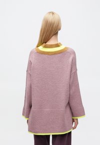 Világos rózsaszín kötött pulóver széles, texturált sárga és barna csíkos gallérral, valamint neonzöld szegéllyel az alján és az ujjaknál.