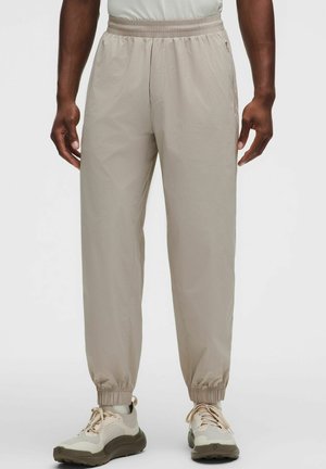 Mann trägt beige elastische Jogginghosen mit Bund und Knöcheln sowie beige-weiße Sneakers vor einfarbigem Hintergrund.