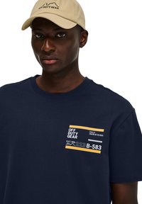 Marineblauwe T-shirt met een gestructureerd ontwerp, voorzien van een witte en gele grafiek op de linkerborst. Een beige pet maakt de look compleet.