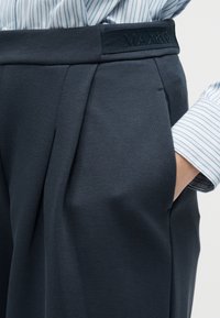 Κοντινό πλάνο σε σκούρο ναυτικό παντελόνι με πιέτες, λάστιχο στη μέση με την ένδειξη «MAX&Co» και χέρι μέσα στην τσέπη, φορεμένο με ριγέ πουκάμισο.