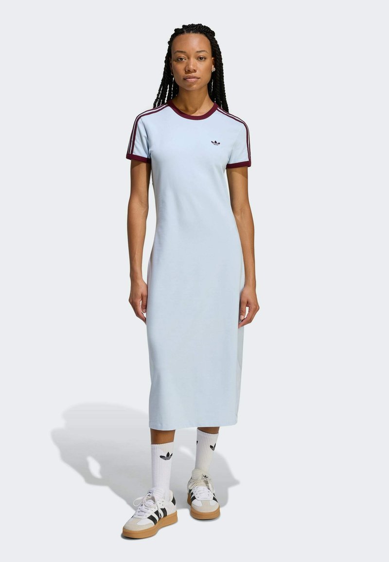 Hellblaues Baumwollkleid mit lockerem Schnitt, kurzen kastanienbraunen Ärmeln und drei weißen Streifen. Model trägt weiße Turnschuhe und Socken mit Logos.