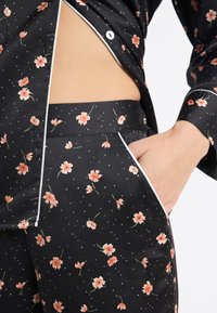 Czarny satynowy komplet piżamowy z rozproszonymi różowymi kwiatowymi wzorami i białymi akcentami. Zawiera koszulę na guziki i dopasowane spodnie z bocznymi kieszeniami.