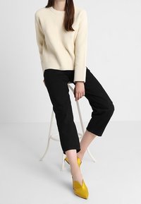 Cremefarbener gerippter Pullover, schwarze cropped Hose und gelbe spitze Schuhe. Das Outfit hat eine entspannte Passform und eine schlichte Textur.