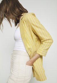 Żółta marynarka w paski wykonana z lekkiego materiału, z podwiniętymi rękawami i luźnym krojem, połączona z białą bluzką i beżową spódnicą.