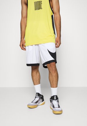 Mann trägt ein gelbes ärmelloses Nike-Shirt, weiße Basketballshorts mit schwarzem Nike-Swoosh, weiße Socken und schwarz-weiße Basketballschuhe.