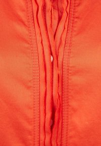 Camicia button-up arancione con pieghe verticali lungo la parte anteriore, con un singolo bottone bianco visibile al centro e una texture di tessuto liscia.