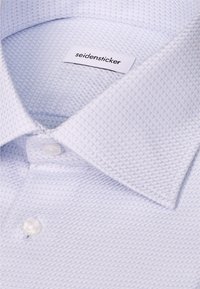 Gros plan sur le col d’une chemise habillée bleu clair texturée avec des boutons blancs et une étiquette de marque Seidensticker à l’intérieur.