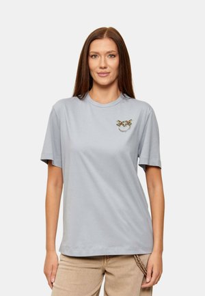 Donna con lunghi capelli castani che indossa una maglietta a maniche corte grigio chiaro con un piccolo emblema colorato sul lato sinistro del petto e pantaloni beige.