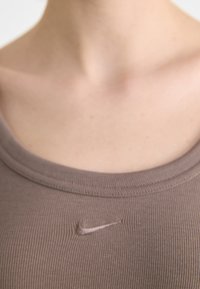 Šviesiai rudas raštuotas liemenė su apvalia iškirpte ir maža Nike logotipu priekyje. Audinys atrodo minkštas ir prigludęs.