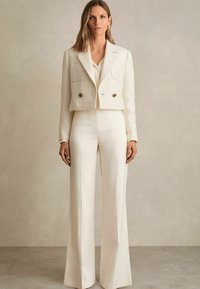 Moteris, aps wearing cream spalvos šviesiai ruda trumpa švarka su auksinėmis sagomis ir plačiomis aukštai krypstančiomis keliantėmis kelnėmis, stovi prieš neutralų foną.