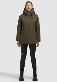 Brown insulated jacket with a high collar and hood, featuring zippered side pockets, paired with black leggings and combat boots.