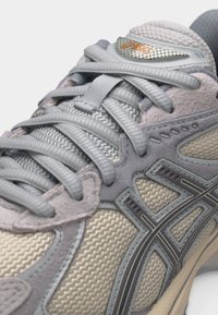 Zbliżenie na szarobeżowy but sportowy ASICS z wzorzystą siateczką, zamszowymi nakładkami i zawiązanymi sznurówkami na białym tle.