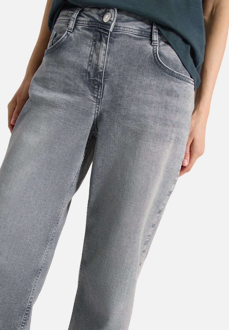 Personne portant un jean en denim gris délavé avec une fermeture boutonnée, des poches avant et un haut sombre rentré.