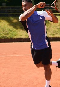 Blauw en wit sportshirt met zwarte accenten, gecombineerd met zwarte structuurshorts; speler slaat een tennisbal op een oranje gravelbaan.