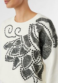 Biały sweter z misterną czarną haftowaną dekoracją liści i abstrakcyjnych kształtów, z różnorodnymi fakturami i wzorami na całej powierzchni.