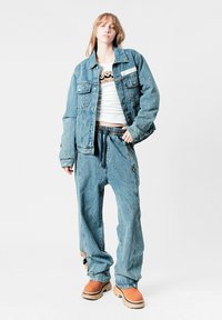 Chaqueta de mezclilla y jeans holgados en azul claro, combinados con una camisa blanca con un diseño estampado. Botas marrones con suelas gruesas.