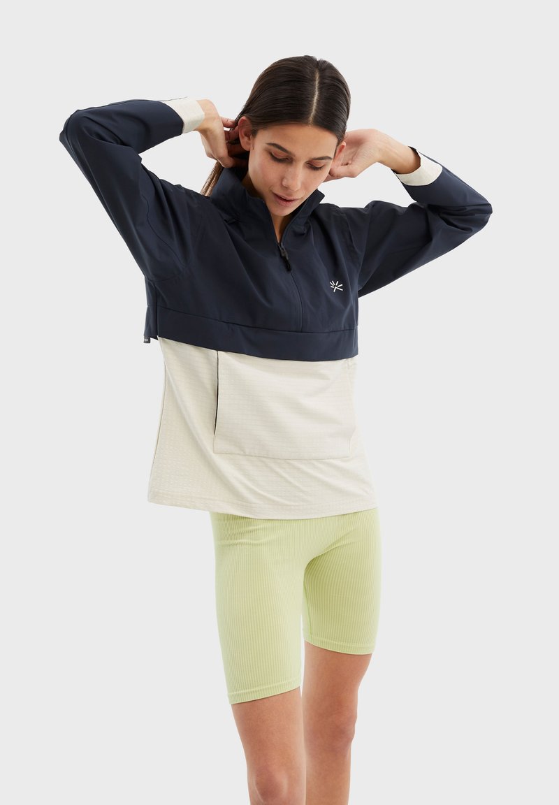 Marinblå och krämfärgad lätt jacka med färgblockering och ficka fram, i kombination med ribbade ljusgröna cykelshorts. Har en dragkedja vid kragen.