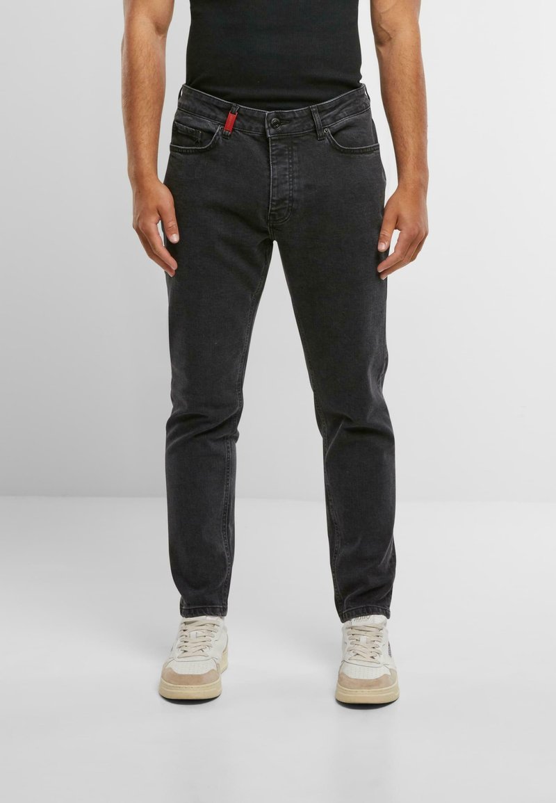 Vaqueros de mezclilla negra de corte slim, con bolsillos estándar y un acento rojo en la parte frontal. Combinados con zapatillas de color claro.