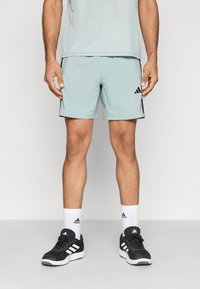 Αθλητικό σορτς ανοιχτού μπλε χρώματος με λεία υφή, διακοσμημένο με μαύρες πλευρικές λεπτομέρειες και λογότυπο Adidas. Συνδυάζεται με μαύρα παπούτσια και λευκές κάλτσες.