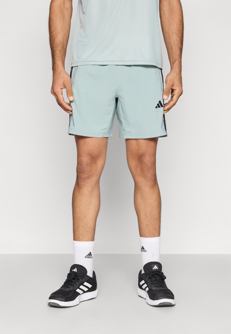 Αθλητικό σορτς ανοιχτού μπλε χρώματος με λεία υφή, διακοσμημένο με μαύρες πλευρικές λεπτομέρειες και λογότυπο Adidas. Συνδυάζεται με μαύρα παπούτσια και λευκές κάλτσες.